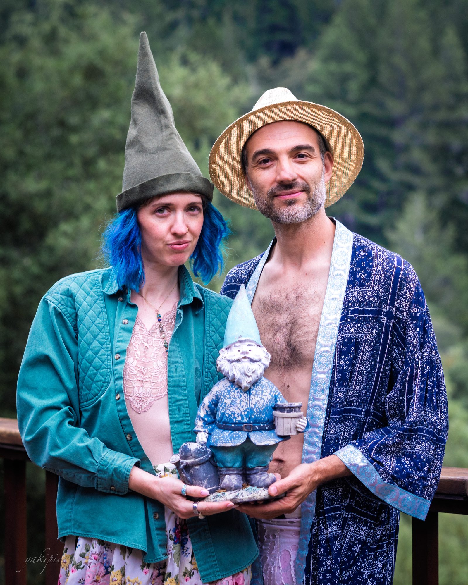 Playfully dressed couple holding a garden gnome in a wooded setting.