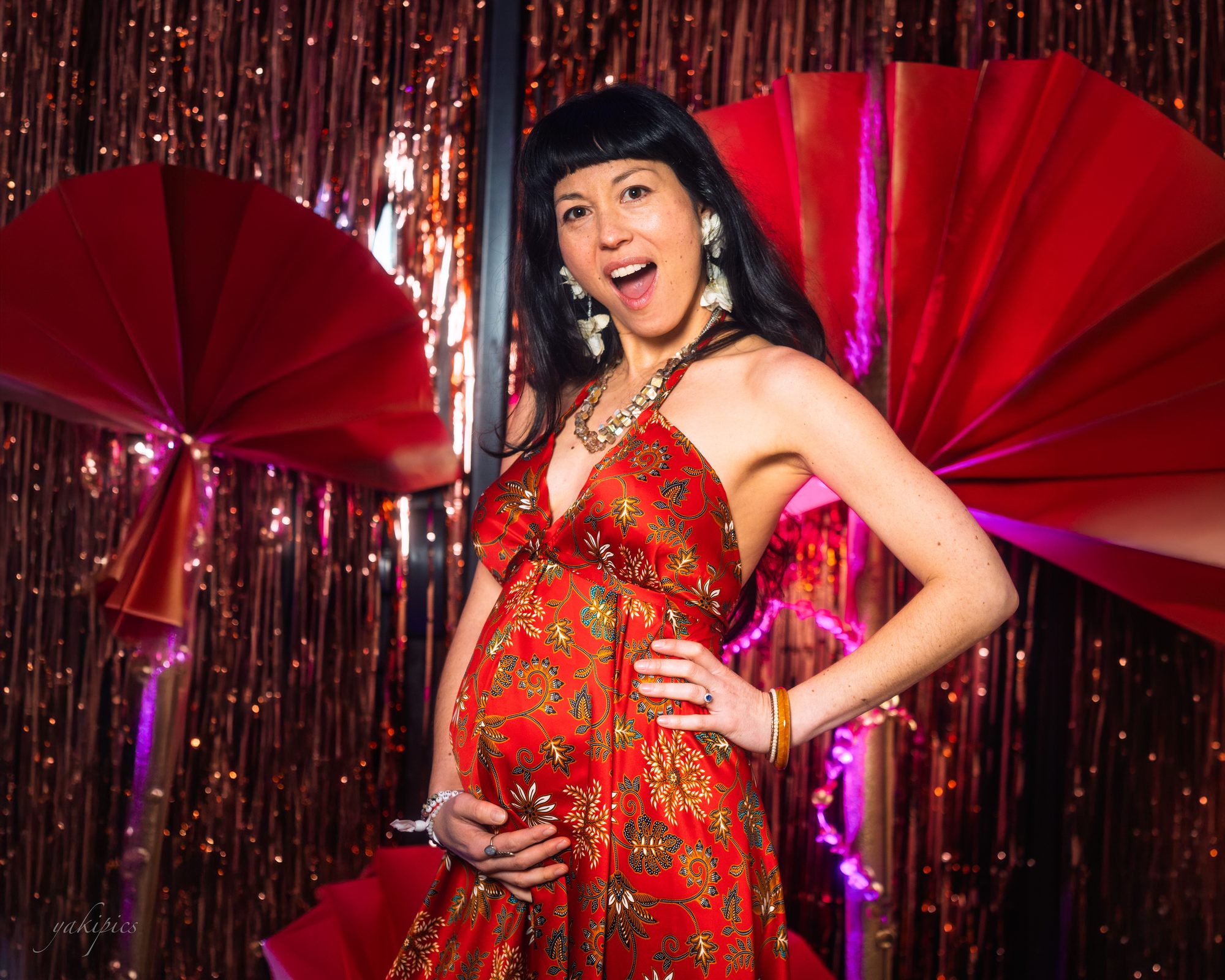 Pregnant woman in a patterned red dress posing exuberantly among red fans and tinsel.