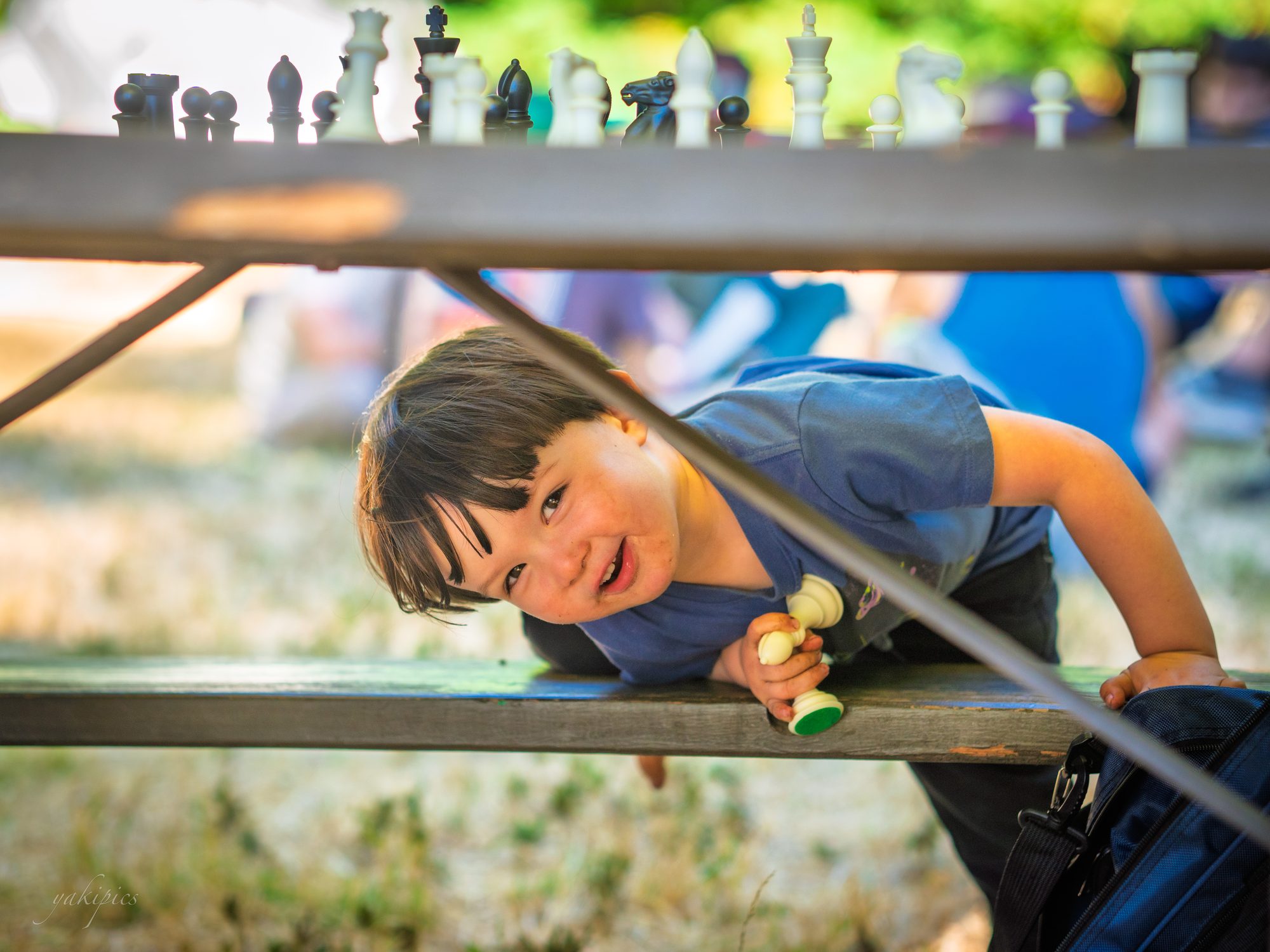Young child peeking sideways from beneath a table lined with chess pieces.