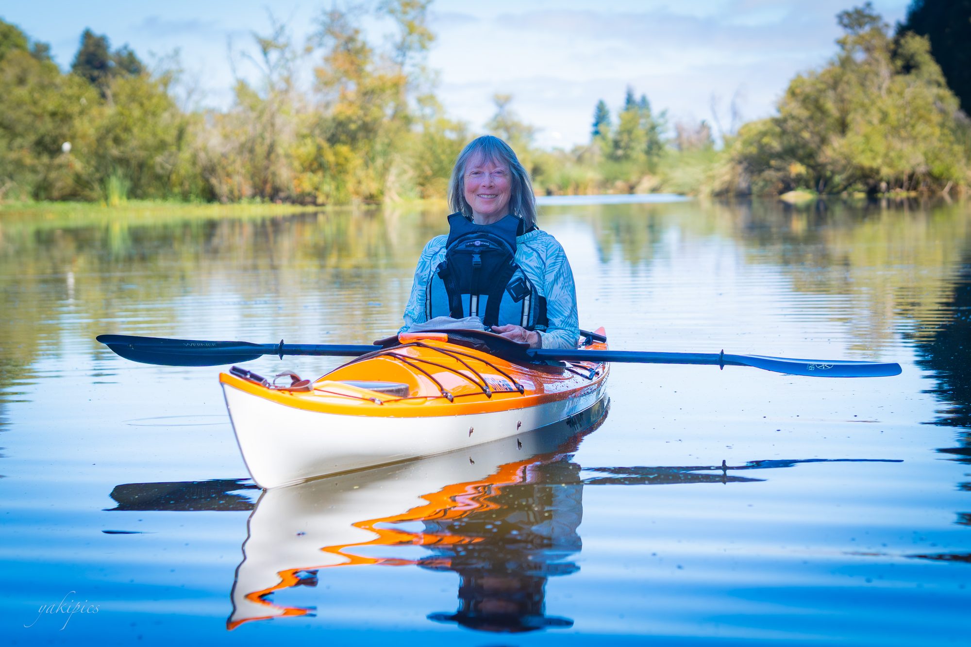 Woman smiling from an orange kayak on a calm, tree-lined river.