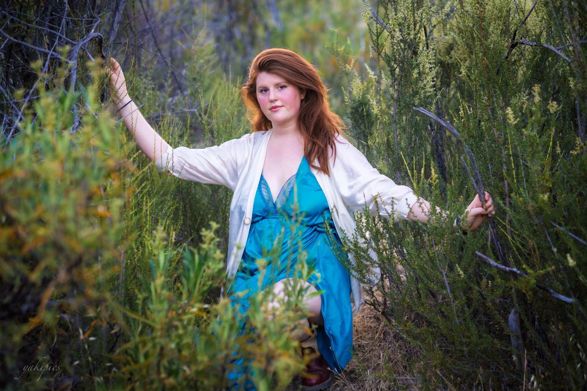 Red-haired woman in a blue dress standing among dense coastal brush.